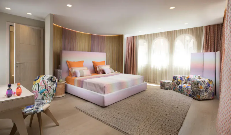 Luxurious Byblos Palace hotel bedroom in Saint-Tropez with pink bed, colorful pillows, patterned chairs, and arched windows.