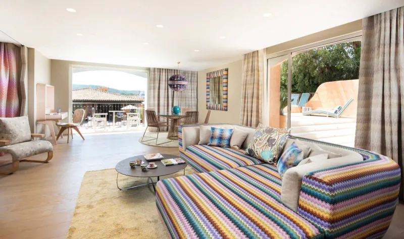 Spacious luxury hotel living room with colorful striped L-shaped sofa, balcony view of Saint-Tropez hills.