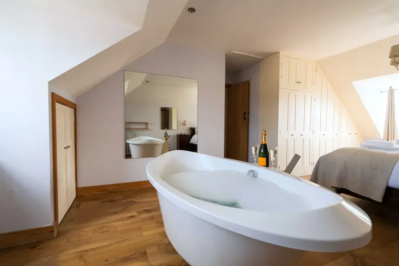 Elegant attic bathroom in Cotswolds country house with freestanding tub, champagne, and bedroom view.