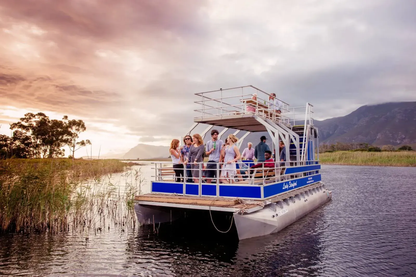 Group of smiling people on upper deck of blue Lady Stanford cruise boat on Klein River at sunset, mountains backdrop.