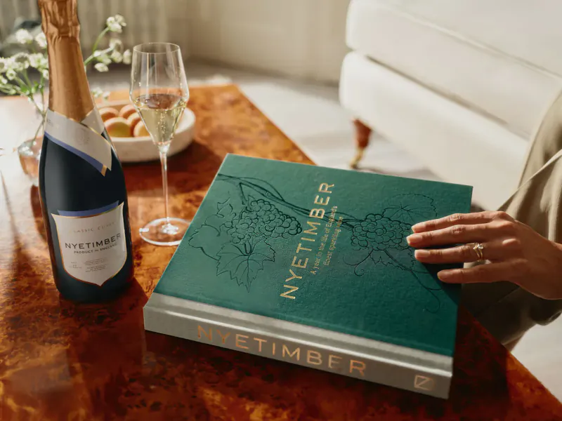 Nyetimber champagne bottle, flute of sparkling wine, macarons on wooden table with open green Nyetimber book, hand turning page.