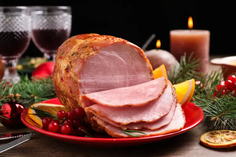 Sliced glazed ham on red plate with Christmas decorations, cranberries, oranges, wine glasses, and candles.