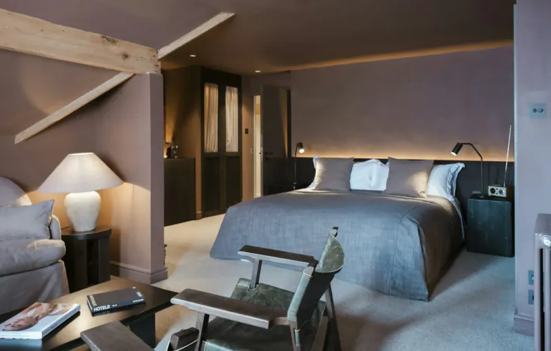 Luxurious hotel suite with king bed, armchairs, lamps, and exposed beams in warm lighting.