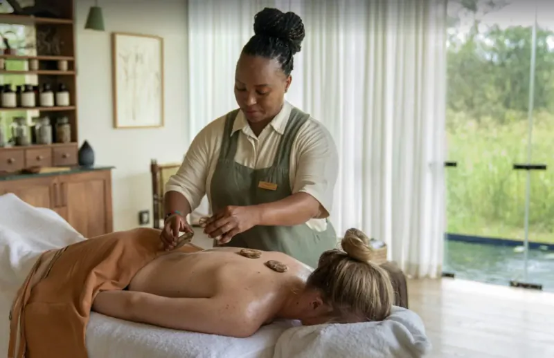 Therapist placing hot stones on client's bare back during spa treatment at eco-lodge with bushveld view.