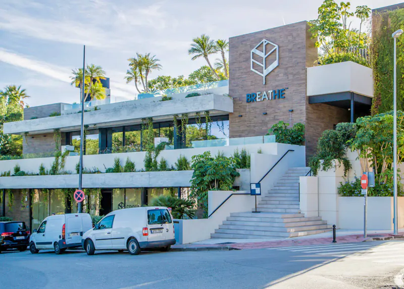 Breathe Marbella modern building exterior with 'Breathe' logo, palm trees, plants, stairs, and parked vans.