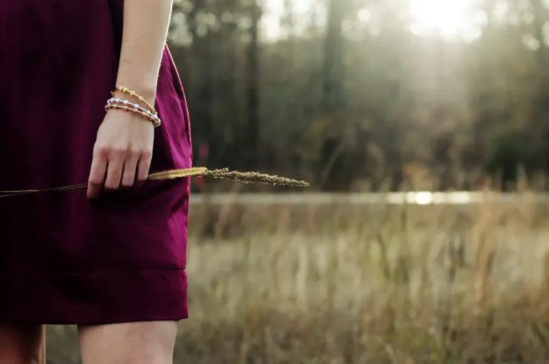 Woman in maroon dress holding golden wheat stalk in sunlit field, wearing beaded bracelets