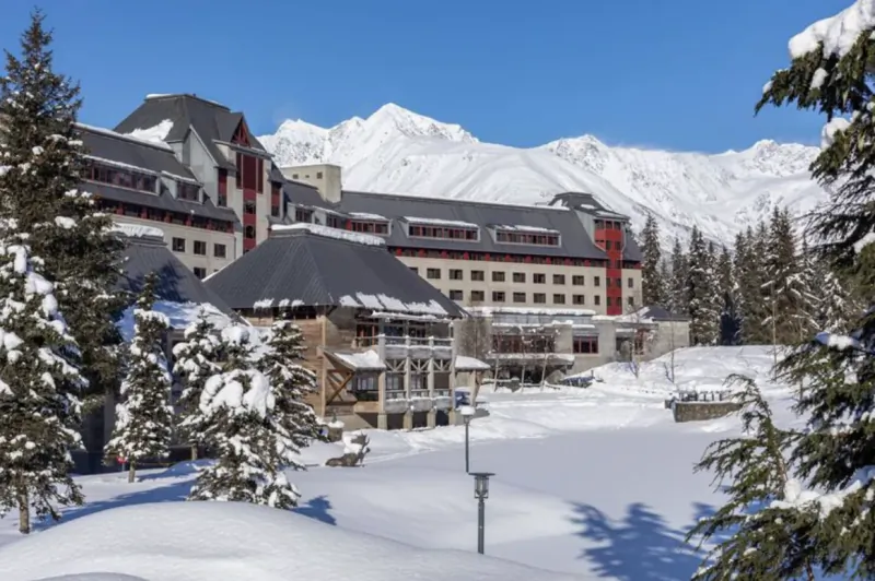 Luxury ski resort with snow-capped mountains, evergreen trees, and blue sky backdrop.