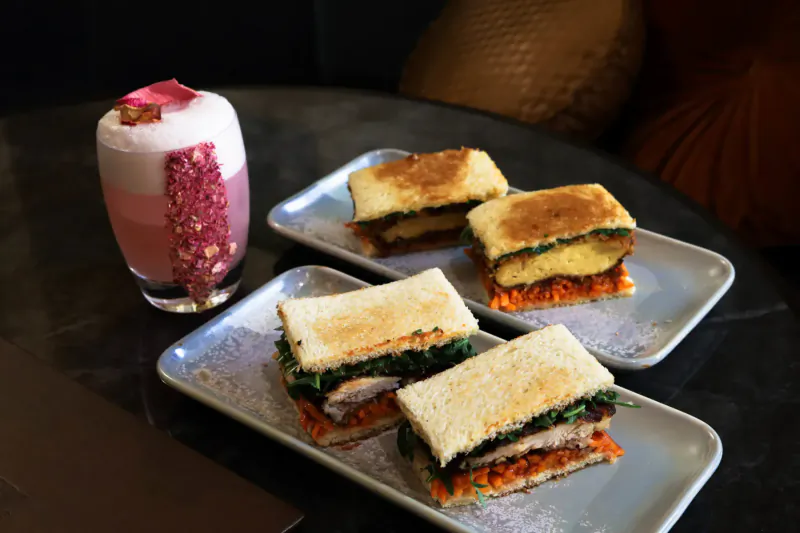 Plated gourmet sandwiches with chicken, greens, carrots on dark table beside pink rose foam drink