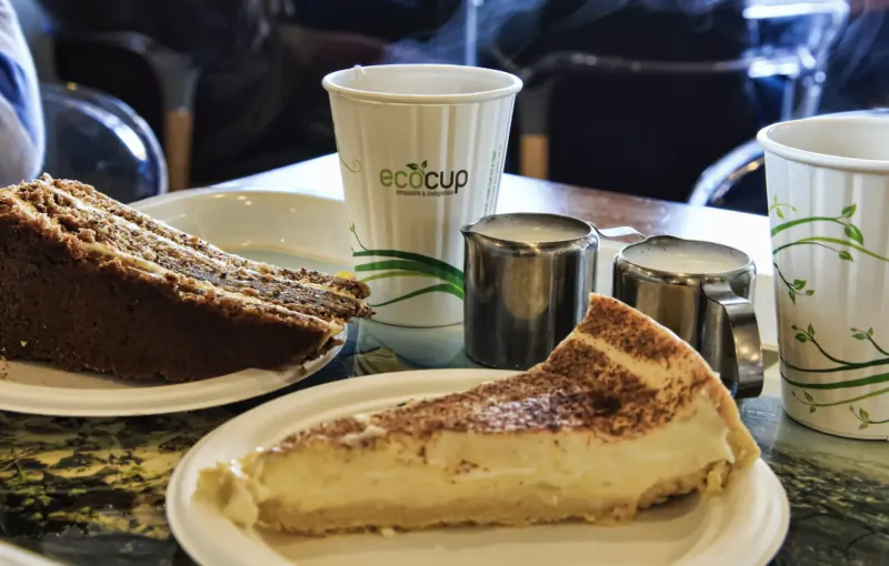 Ecocup branded cups, salt and pepper shakers, layered cake, and cheesecake on plates at cafe table.