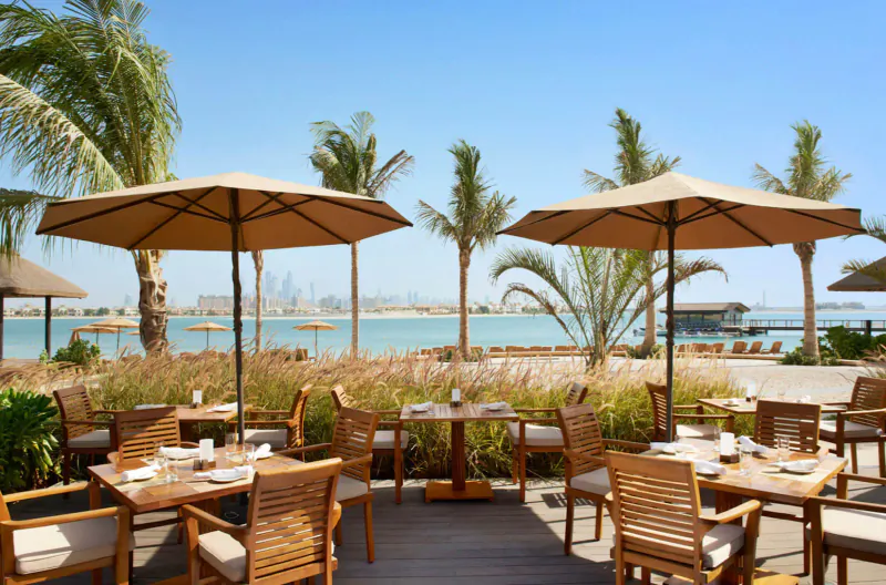 Outdoor dining area at Sofitel Dubai the Palm with wooden tables, beige umbrellas, palm trees, and Dubai skyline over lagoon under blue sky.