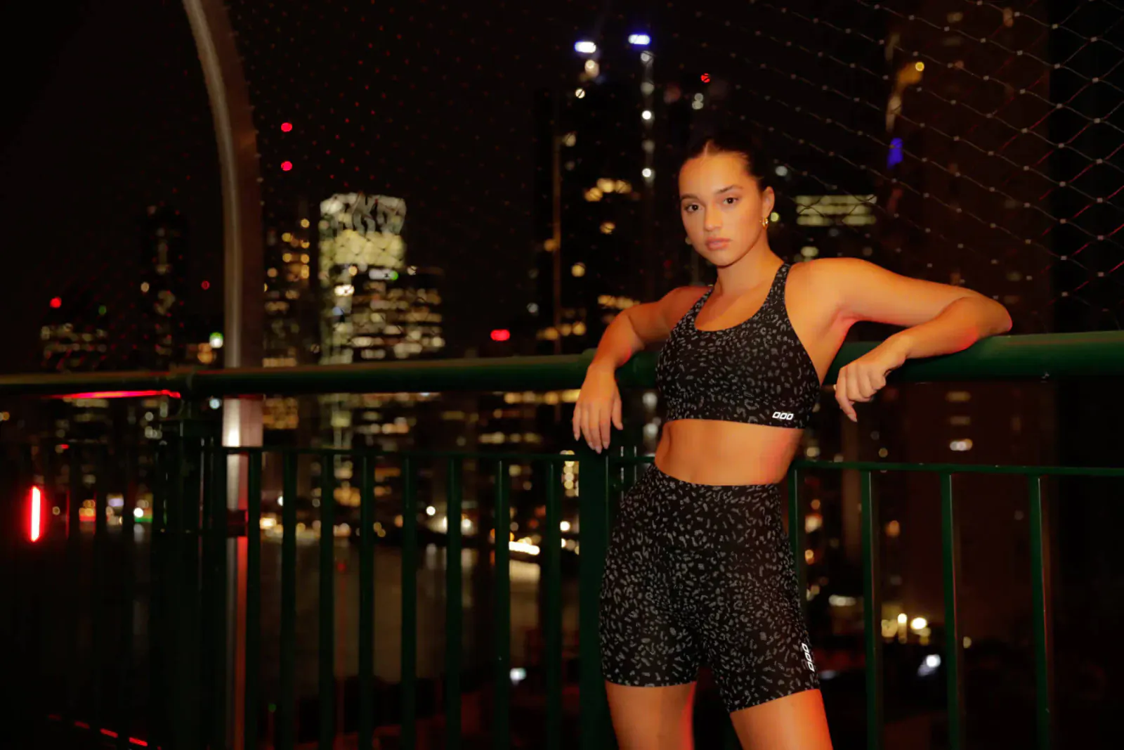Young woman in black Lorna Jane leopard-print activewear leans on green railing at night with city skyline.