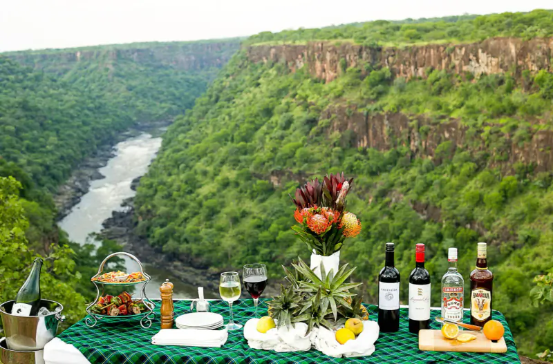 Riverside picnic table at Old Drift Lodge with wine bottles, fruits, cheese, and river gorge view.