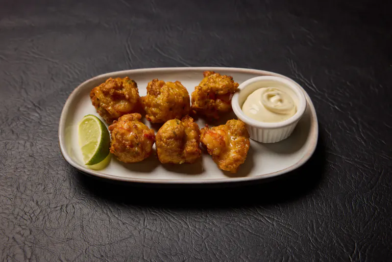 Six crispy battered shrimp fritters with lime wedge and white dipping sauce on oval plate