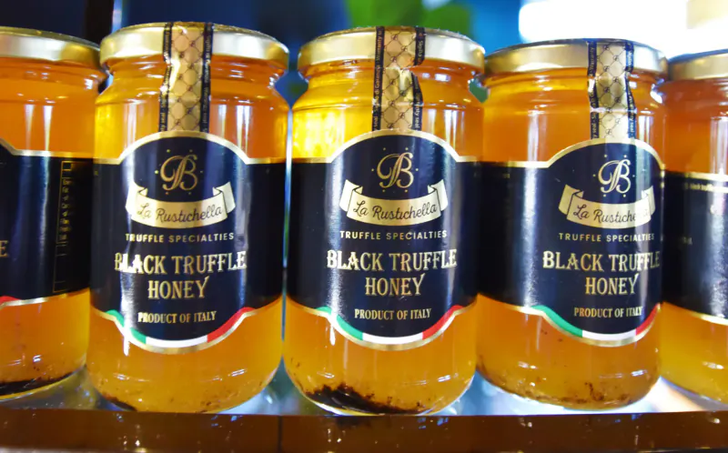 Row of La Bottega black truffle honey jars from Italy on shelf, golden honey with truffle bits visible.