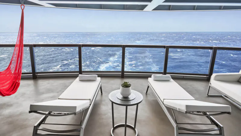 Luxury adults-only cruise balcony with red hammock, white loungers, plant table, and ocean view.