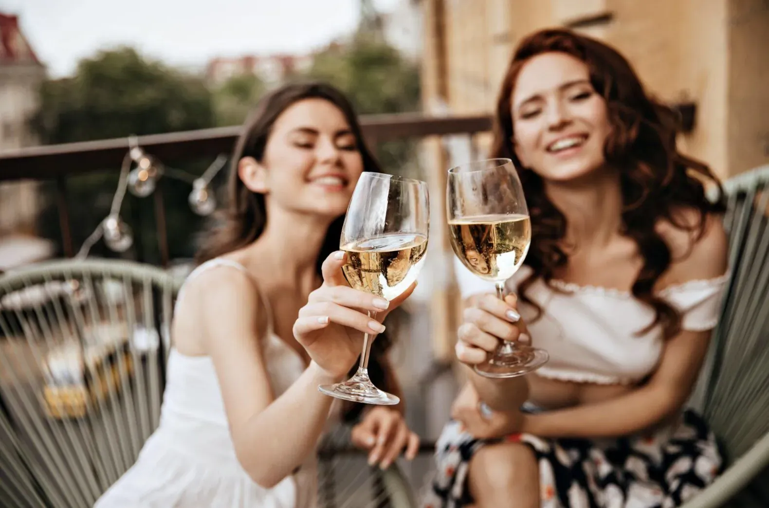 Two smiling women in white dresses clinking glasses of white wine on a balcony with string lights, evoking French elegance.
