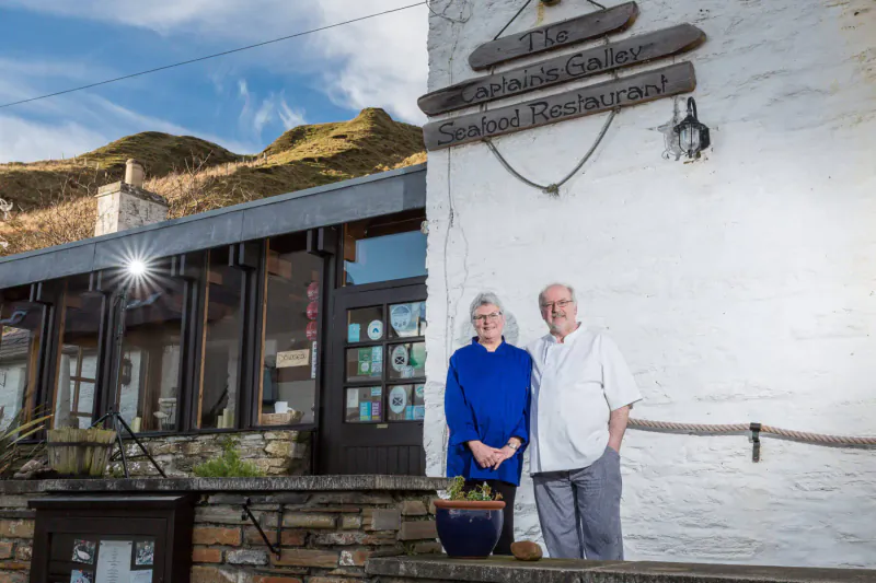 Elderly couple smiling in front of Glen Gallery Seafood Restaurant, white stone building with mountains behind.