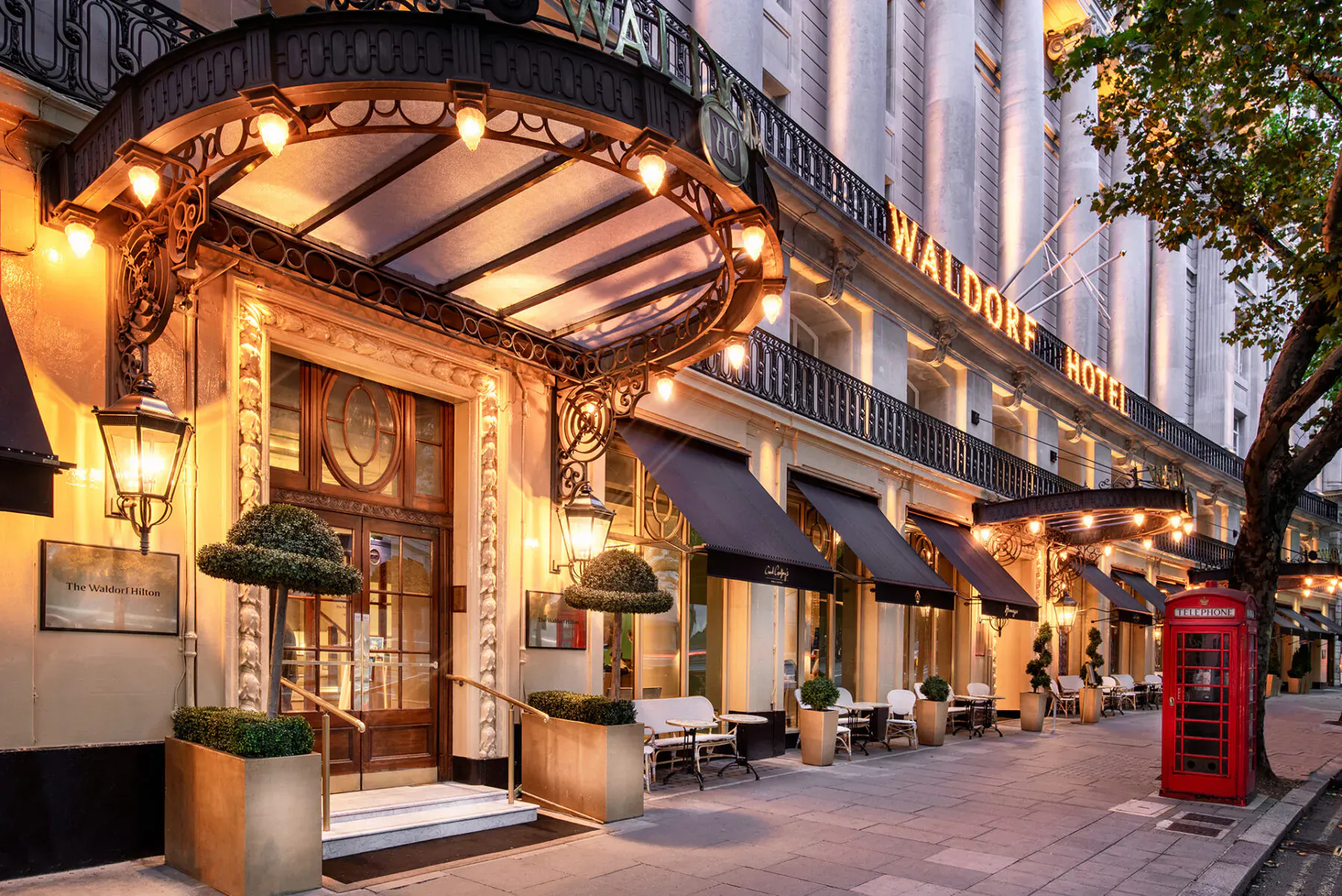 The Waldorf Hilton hotel entrance in London at dusk, with ornate canopy, lanterns, plants, chairs, and red phone booth.