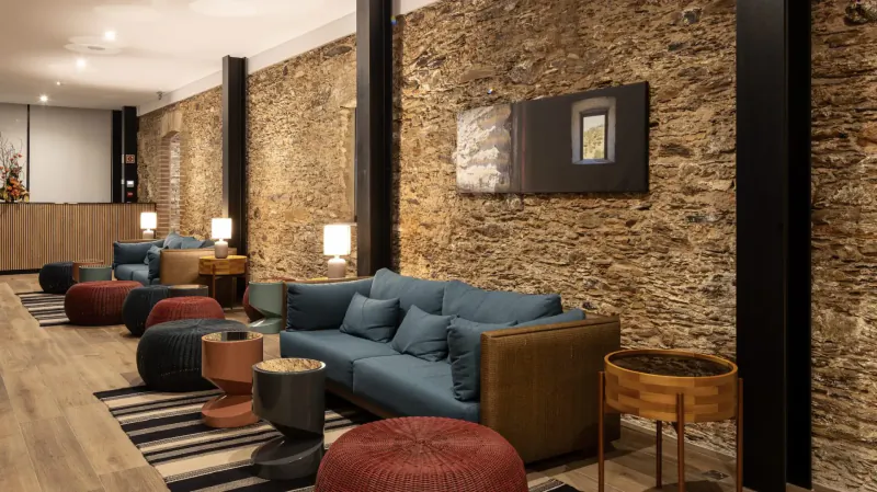 Modern lounge with teal sofas, colorful poufs, wood tables against brick wall and black pillars.