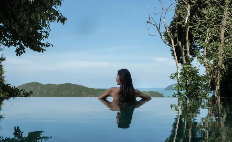 Woman with long dark hair sits topless in infinity pool at Ambong-Ambong, Langkawi, with lush trees, mountains, and sea view.