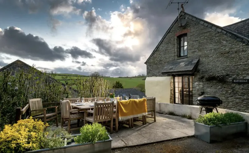 Stone luxury cottage patio with wooden dining table set with yellow cloth, chairs, BBQ, plants, and countryside view at sunset.