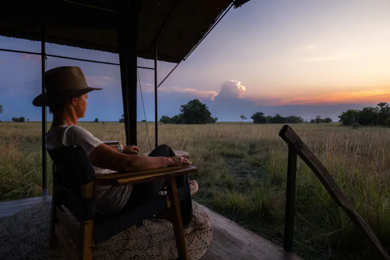 Woman in hat sits pensively on safari deck at dusk, overlooking Zambian savanna grassland.