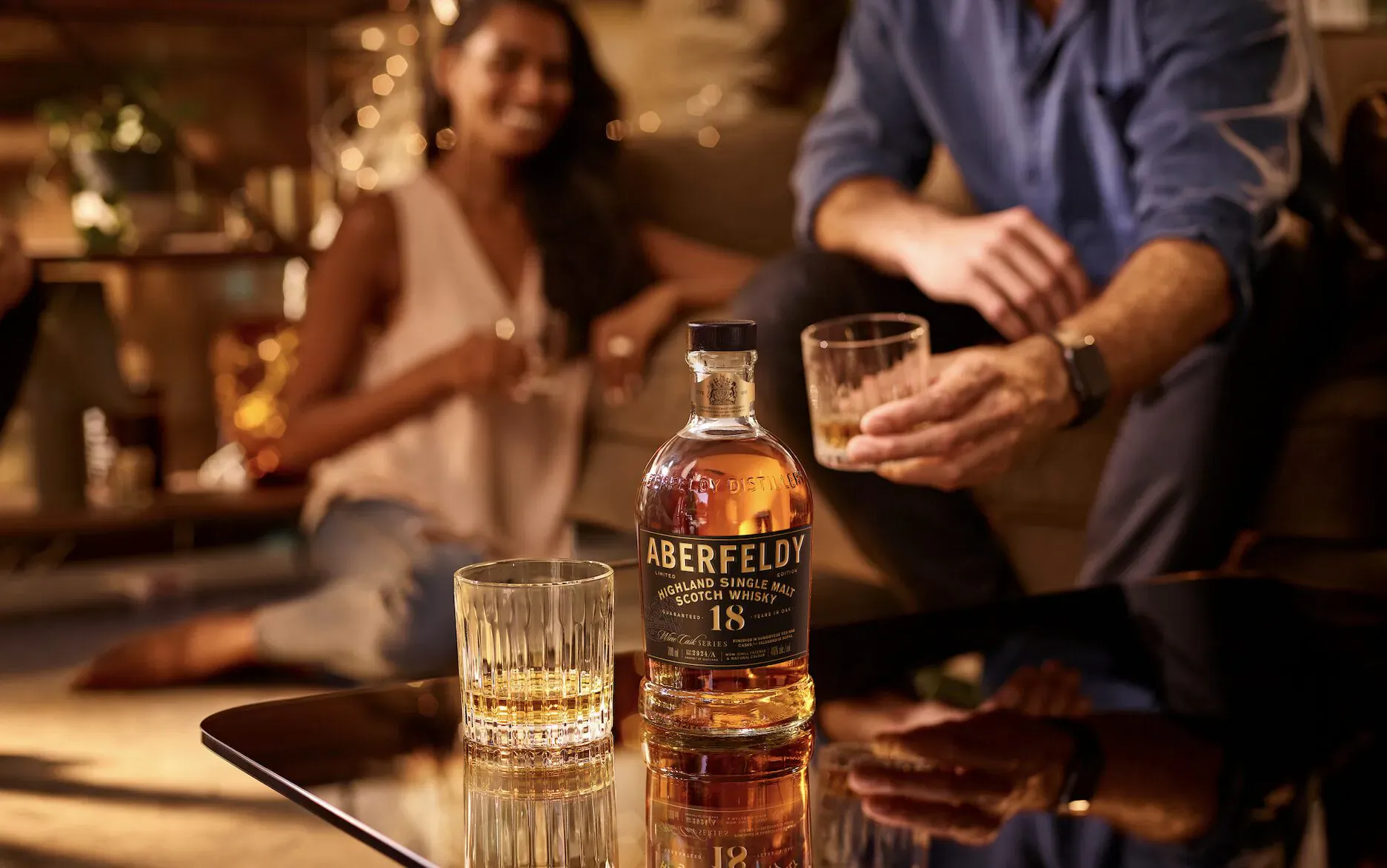 Close-up of Aberfeldy 12 whisky bottle and glasses held by a man and woman in warm indoor setting