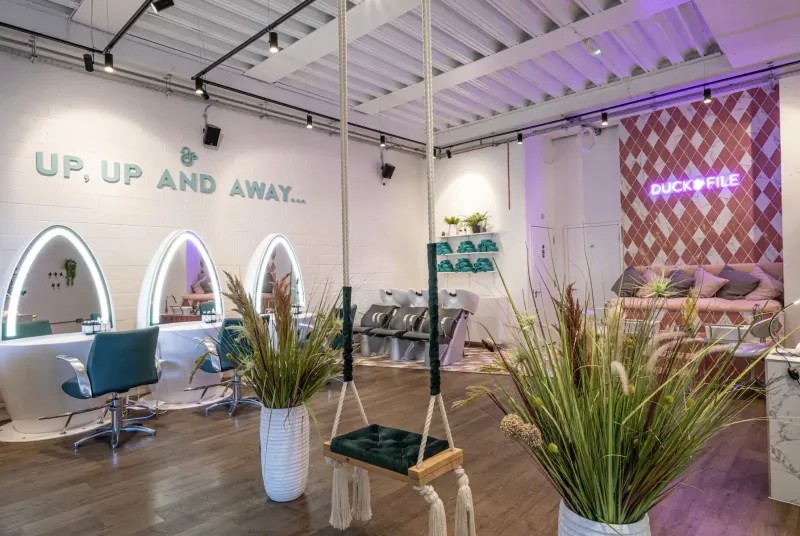Modern Duck & Dry salon interior with 'UP, UP AND AWAY' neon sign, swing chairs, teal styling stations, and potted plants.