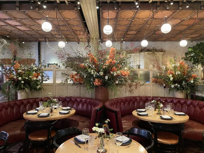Elegant restaurant interior with red curved booth, round wooden tables set for dining, large orange flower arrangements, and exposed beamed ceiling.