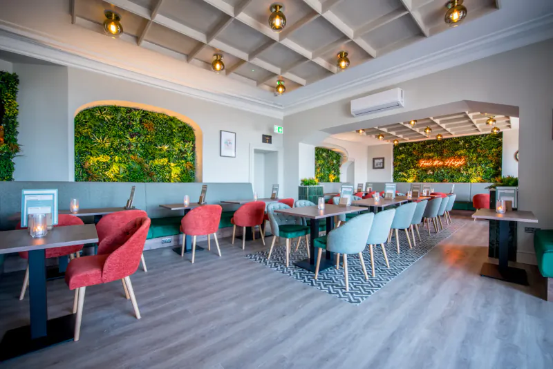 Modern restaurant interior at The Imperial Hotel in Tenby with green wall gardens, red and teal chairs, wooden tables, and pendant lights.