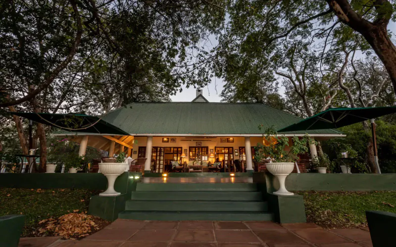 The River Club gallery in Livingstone, Zambia: green-roofed building with verandas, potted plants, steps, and trees at dusk.