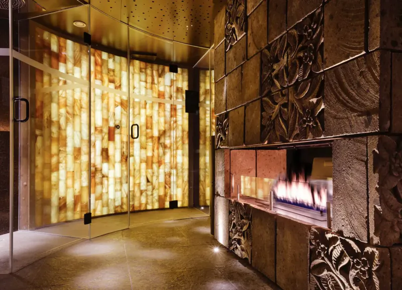 Luxurious spa entrance with onyx glass doors and ornate stone walls beside a glowing fireplace