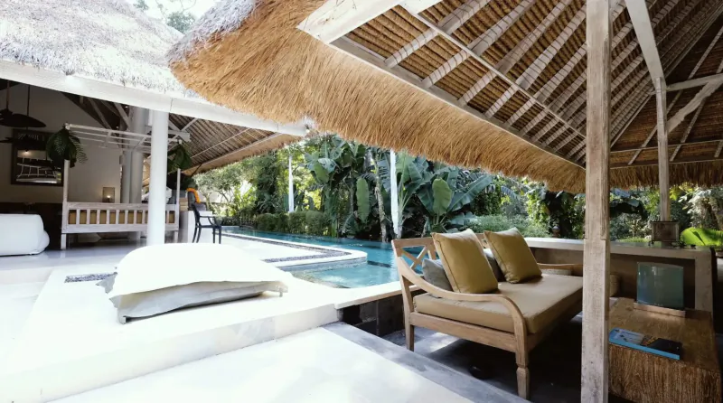 Open-air Bali villa with thatched roof, white daybed, teak sofa by infinity pool in lush jungle setting