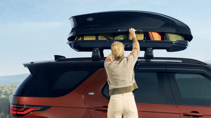 Woman loading gear into black roof box on red Land Rover Discovery SUV in scenic outdoors