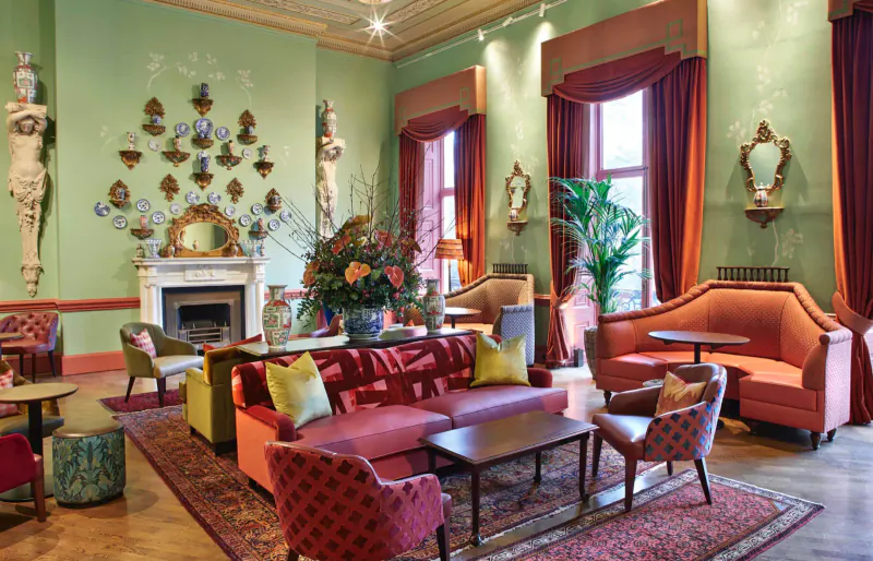 Elegant green lounge at Home House club with red sofas, ornate fireplace, statues, and large floral arrangement.