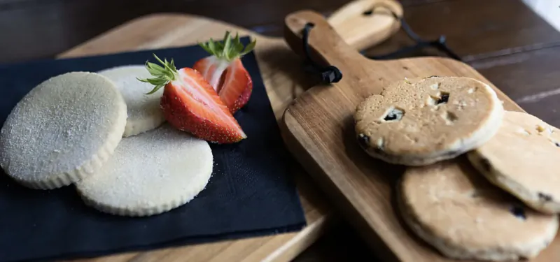 White shortbread cookies with fresh strawberries on black plate beside chocolate chip pancakes on wooden board