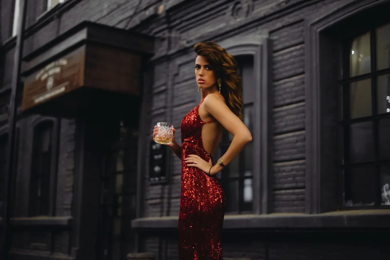 Brunette woman in sparkling red backless dress holding cocktail, posing sideways outside dark brick building
