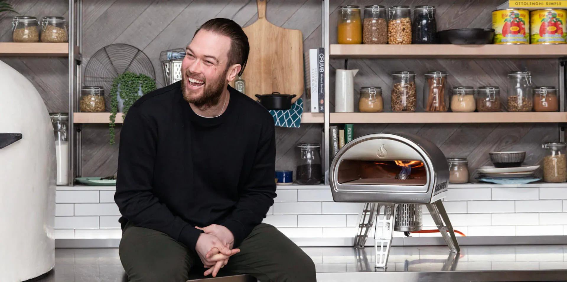 Tom Gozney smiling in modern kitchen beside lit Gozney pizza oven on shelves with jars
