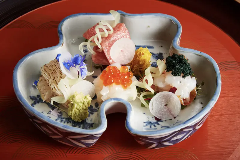 Kaiseki dish with salmon roe, pink radish, blue flowers, and greens on blue clover-shaped plate on red tray.