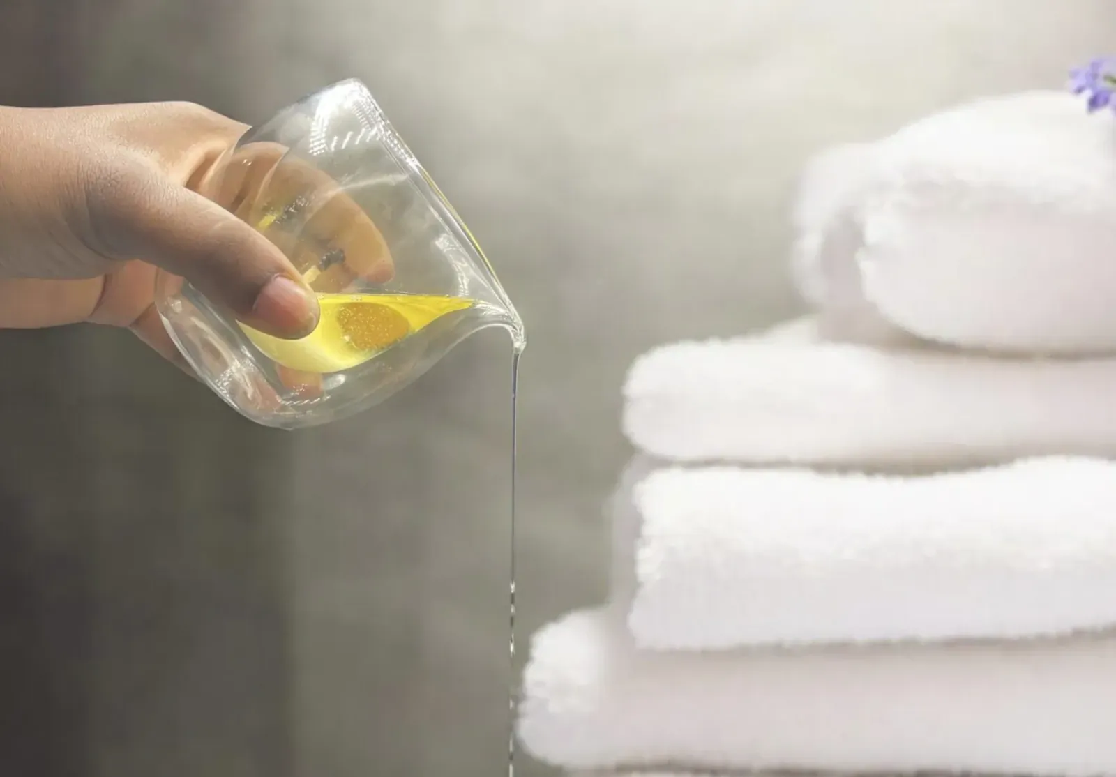 Hand pouring yellow amber liquid from glass cup onto stacked white towels with purple flowers
