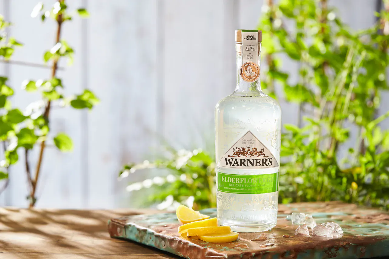 Warner’s Gin bottle with green label on wooden surface with lemon slices, surrounded by lush green foliage