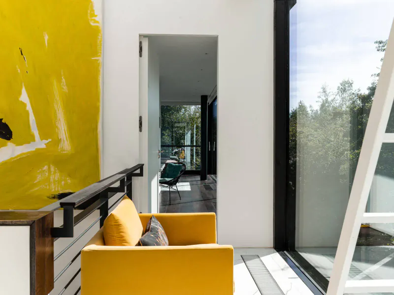 Modern interior of Glasshouse by the Lake gallery: yellow abstract painting, orange chair, white walls, large glass windows to trees.