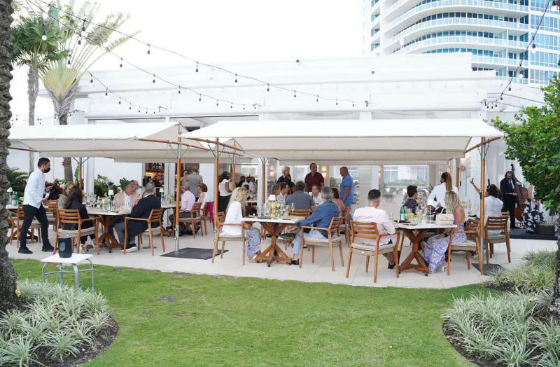 Diverse group dining at outdoor Italian affair under white canopies and string lights, luxury high-rise and palms in background, South Beach wine & food festival.