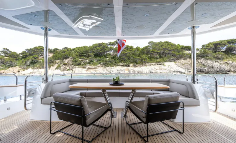 Luxury yacht deck with wooden dining table, cushioned benches, chairs, potted plant, and Union Jack flag overlooking pines and sea.