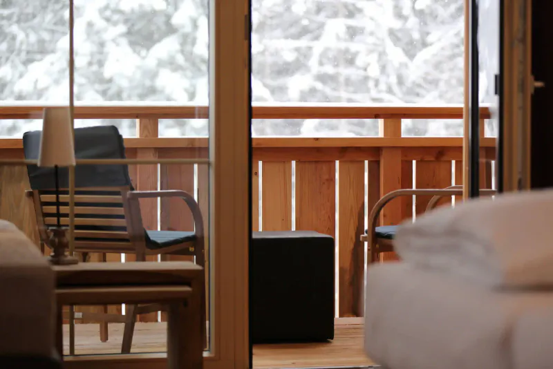 Cozy hotel room with armchair, lamp, ottoman, and bed, overlooking snowy mountain balcony view.