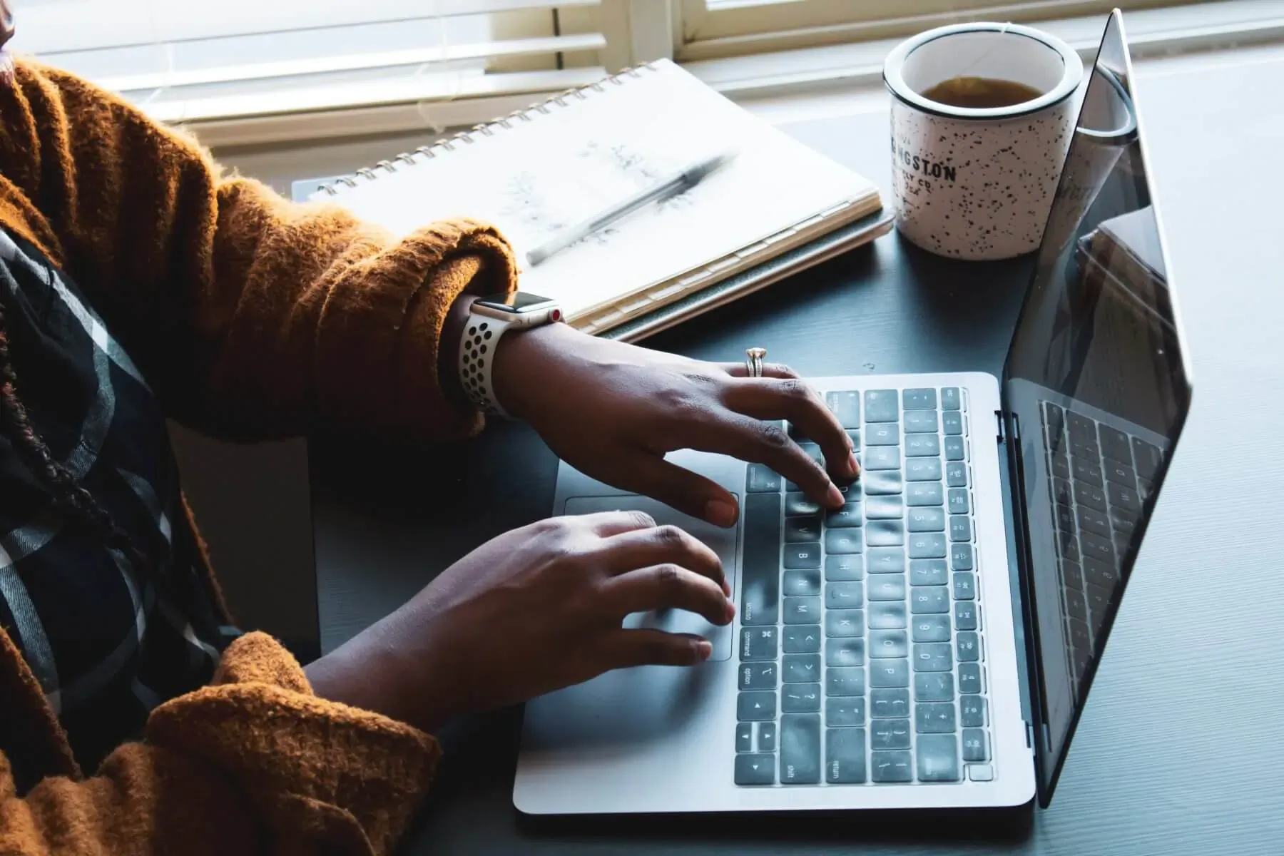 Person typing on MacBook laptop at desk with notebook and coffee mug by window