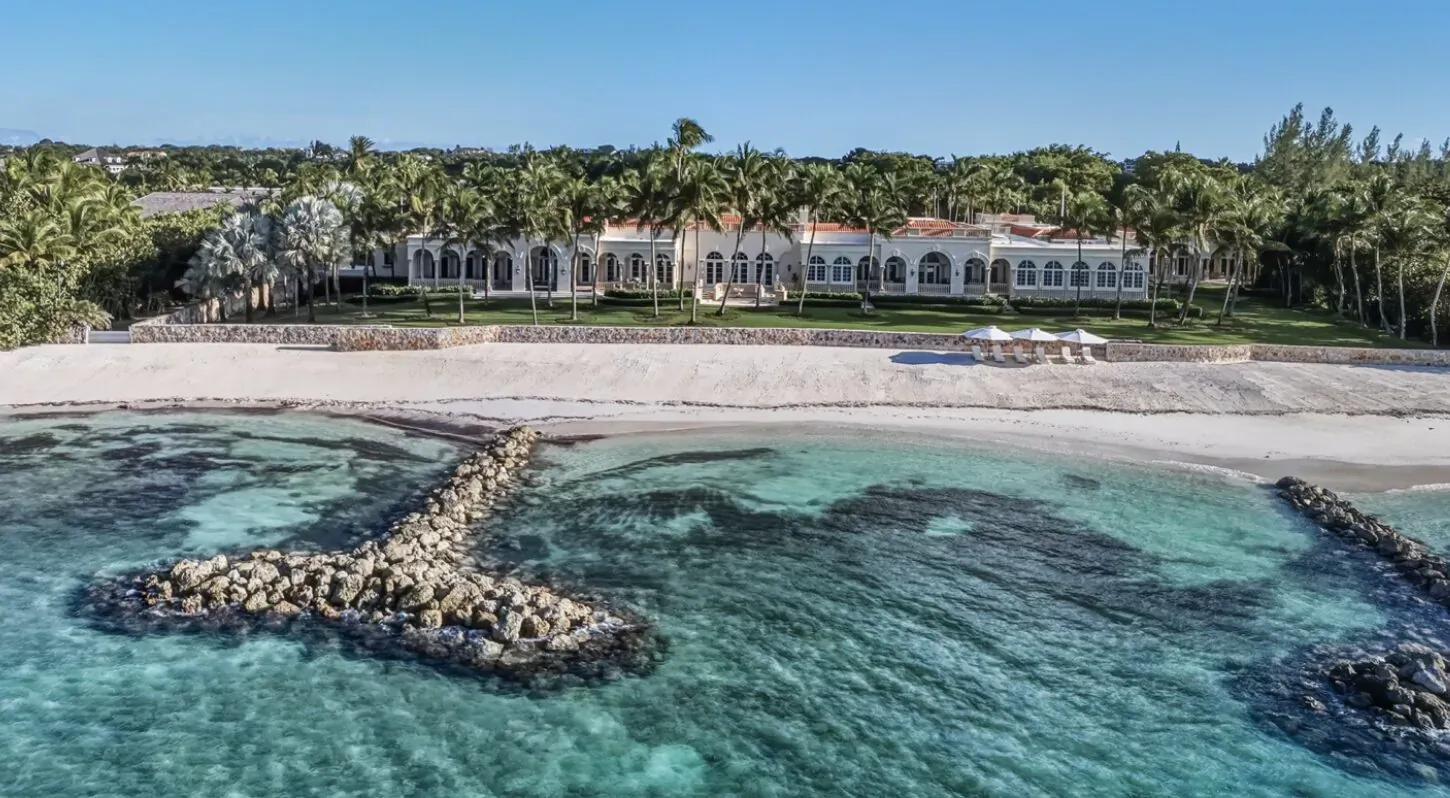 Aerial view of Sans Souci, landmark 5.5-acre oceanfront estate in Lyford Cay, white mansion with arches amid palms on sandy beach with turquoise waters and breakwaters.