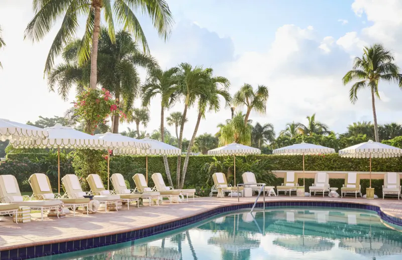 Luxury Palm Beaches poolside with white umbrellas, lounge chairs, palm trees, and blue water under partly cloudy sky.