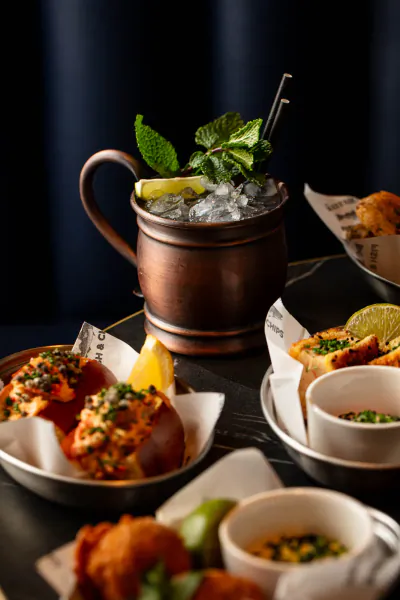 Copper Moscow Mule mug with lime and mint beside lobster rolls, fried bites, and dips on black table at Lobster Pot Booth.