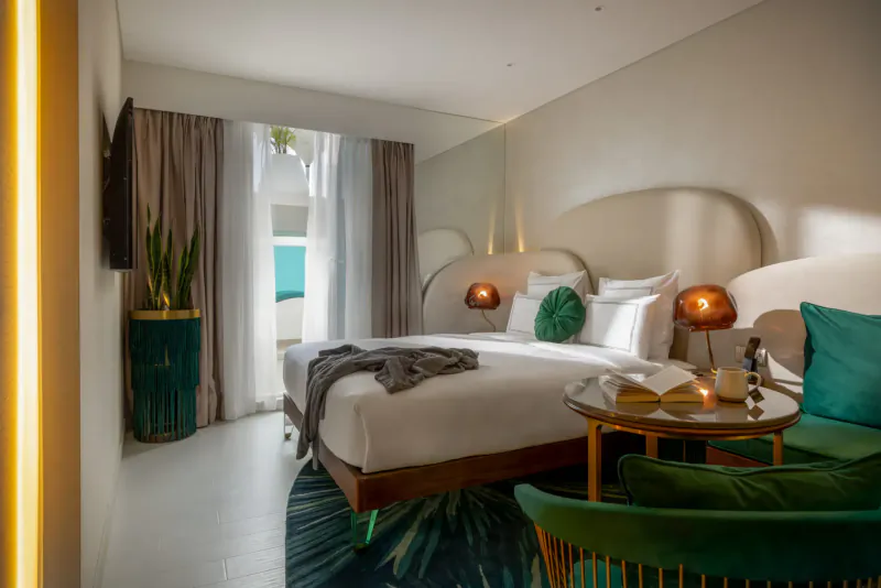 Modern hotel bedroom with curved white headboard, green pillows, bed lamp, side table, green velvet chair, and ocean view balcony.